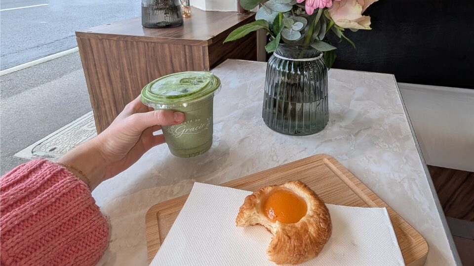 A hand holding a matcha latte and a fruit Danish on a chopping board with a bite taken out of it