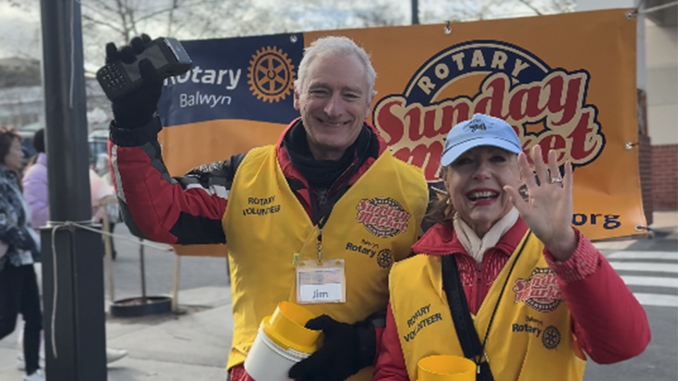 Image of 2 volunteers waving to the camera while standing in front of a hung banner that says Rotary Sunday Market, holding collection buckets for donations.