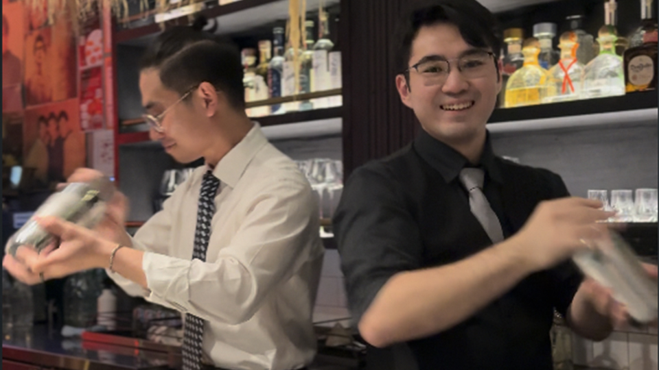 Two bartenders standing behind a bar, in front of a wall of bottles shaking cocktail tins smiling at the camera.