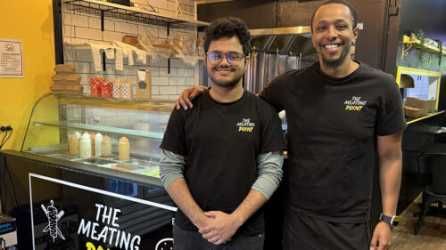 Image of 2 men working at The Meating Point, smiling, with the taller man having his hand on the shoulder of the other man, smiling next to each other, in front of the counter.