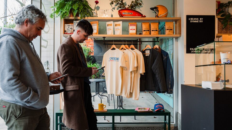 Two customers waiting for coffee in a store with a rack of t-shirts in the background